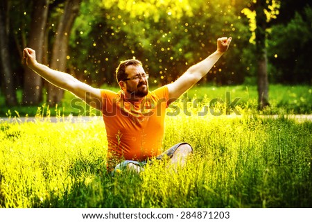 A happy man is relaxing on green grass with squint eyes and raised up to sky arms at sunny summer day at park background. Concept of wellbeing and healthy lifestyle