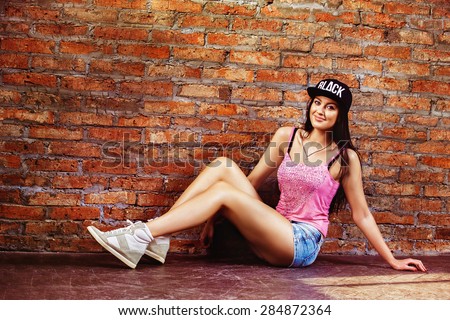 Beautiful young swag style dressed girl in short jeans and cap is sitting at textured red bricks wall background.