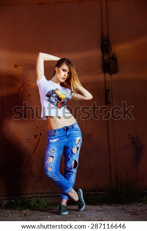 Beautiful young model woman in jeans and handmade shirt is sensually touching hair at red urban rusty gate background.
