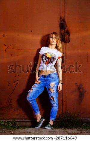 Beautiful young model woman in jeans and handmade shirt is standing at red urban rusty gate background.
