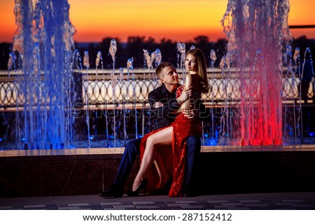 Beautiful young woman in red dress and elegant man sitting on colorful fountain at golden stunning sunset background.