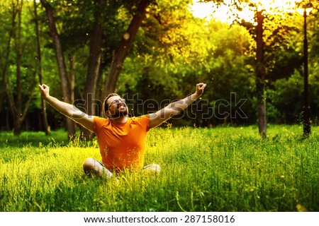 A happy man is sitting on green grass and raising arms up to sky at sunny summer day at park background. Concept of wellbeing and healthy lifestyle