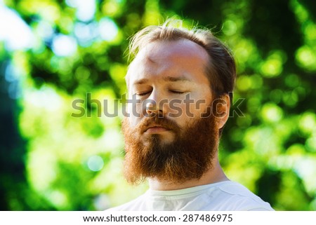 Closeup portrait of man with red beard and closed eyes at green summer park background. Concept of wellbeing, relaxation and meditation.