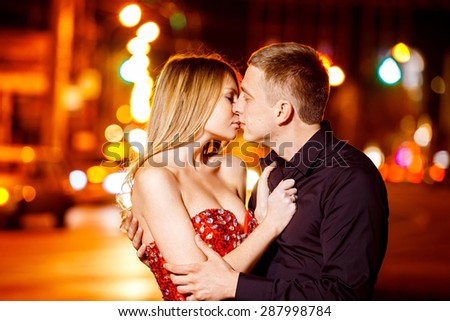 Beautiful young woman in red sparkling dress is passionately kissing elegant macho man at city street at bright lights blurred background.