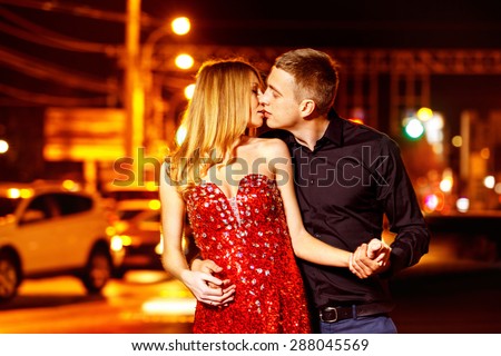 Beautiful young woman in red sparkling dress is passionately kissing elegant macho man at city street at bright lights and moving cars blurred background.