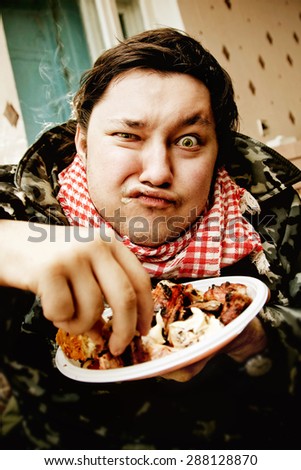 Portrait of greedy hungry chewing man with squint eye taking piece of fat meat from a plate at kitchen background.