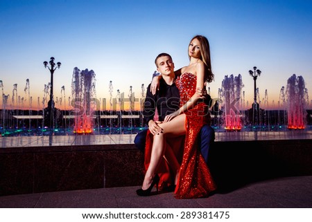 Beautiful young woman in red dress and macho man are sitting at fountain at blue sunset sky background.