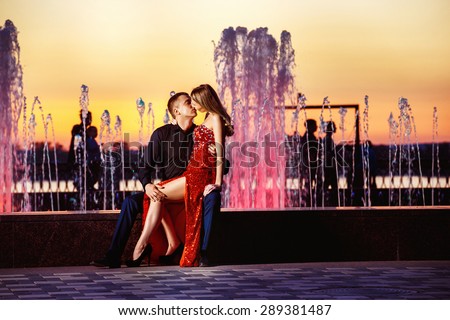 Beautiful young woman in red dress and macho man are kissing at colored water fountain at orange sunset sky background.