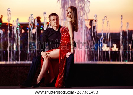 Beautiful young woman in red dress and macho man are sitting on colorful fountain at golden stunning sunset background.