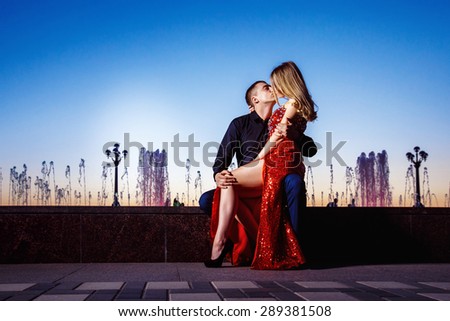 Beautiful young woman in red dress and macho man are kissing at fountain at blue sunset sky background.