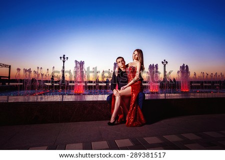 Beautiful young woman in red dress and macho man are sitting at fountain at blue sunset sky background.