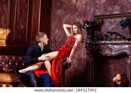 Beautiful young woman in red dress is sitting on sofa and flirting with elegant man in luxury bedroom.