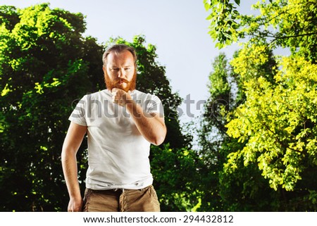 Portrait of serious mature bearded man with red hair touching beard at green summer park background.