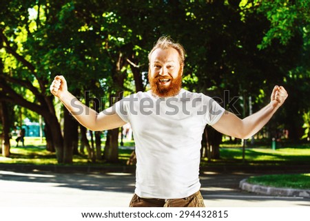 Happy man with red beard is putting hands up as gesture of success, achievement at green summer park background.