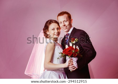 Happy smiling wedding couple holding red roses flowers bouquet at pink wall background.
