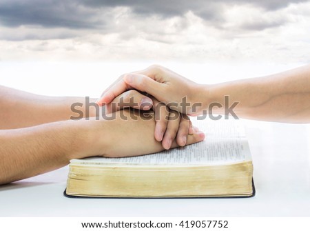 praying hands of two people on burred open bible on white table and burred storm sky  as a background, Christian concept, trust concept