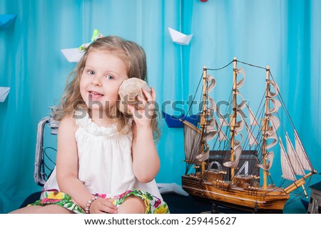 Cute little girl is sitting on a mattress on the sea background scenery, shell keeps hand near ear and smiling