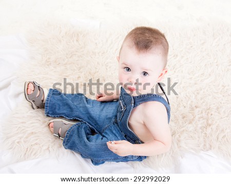 Cute baby with big blue eyes in denim overalls sitting isolated on white background.