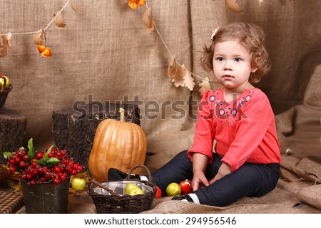Cute curly girl sitting on a background of autumn scenery in the studio. Autumn harvest - pumpkin, apples in boxes. Decorations made of burlap.