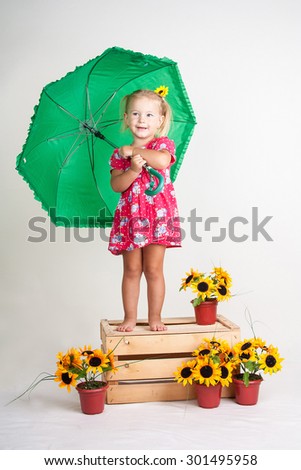 Cute little blond girl standing on a wooden box with a green umbrella in hand. Pots with sunflowers stand near the box. Object isolated on white background