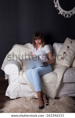 A woman is sitting on a white sofa with a laptop. It corresponds with friends for a chat.