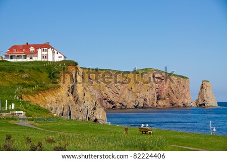 Two people on a bench observing the Red House on the cliff, and part of the  Percé Rock(Percé (city) Quebec, Canada