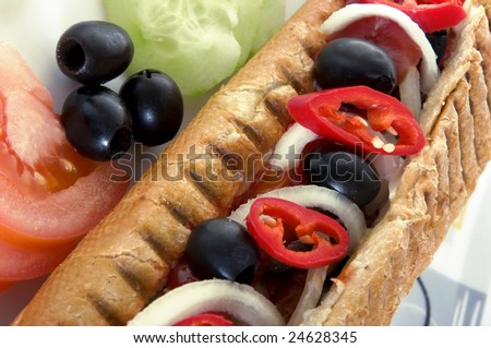 Close-up of hot dog with vegetables. Fast food, tomato, hot pepper, onion, sausage, grilled roll, olives, cucumber.