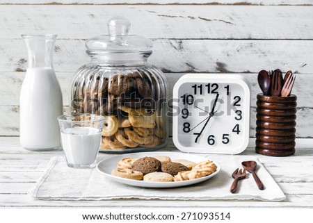 Still life with chocolate chip and butter cookies on white ceramic dish with glass of milk, white clock and wooden spoon on table background