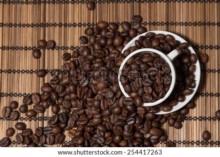 White cup and dish full of coffee beans on bamboo tabletop with spread beans
