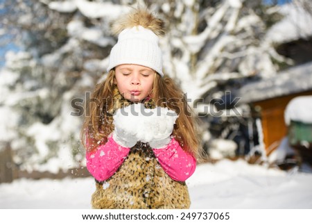 Cute little girl in warm hat blowing snowflakes from hands