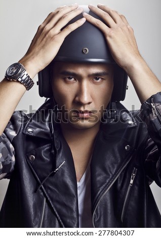 Handsome young Chinese man with black helmet for a motorbike, wearing leather jacket and silver watch, with both hands holding the helmet and looking at camera.
