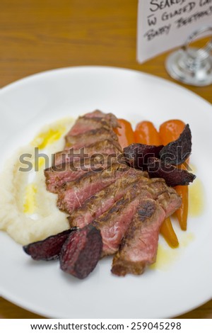 Beef sirloin steak cooked rare, sliced thin and served on white plate with oven roasted vegetables carrot, beetroot and potato glazed with olive oil, and restaurant menu sign.