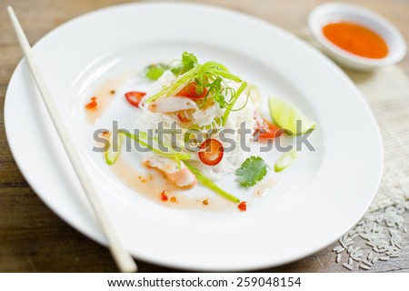 Chili prawn vermicelli noodles garnished with shallots, served with sweet chili sauce and lime. White rice surrounds the white plate which sits on a wooden table.