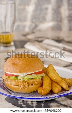Fried chicken, lettuce, tomato and mayonnaise burger served with extra chunky thick chips. Glass of beer in the background to the left and cutlery to the right.