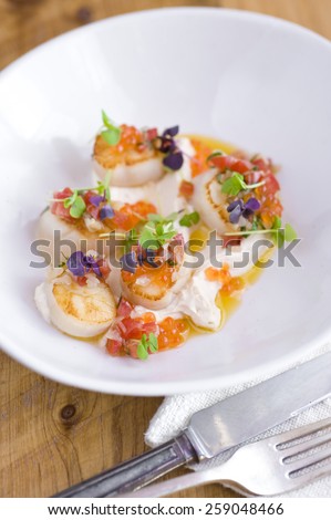 Scallops with their roe off resting in olive oil and thick white sauce/cream cheese/tartar. They are garnished with salmon roe, fine tomato and red onion, and small leaf sprouts. Cutlery bottom right.