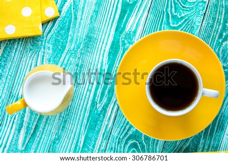 cup of black tea on yellow plate and yellow milk jug\
\
on turquoise colored old wooden table with yellow napkin at polka dots top view