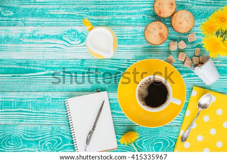 cup of coffee on yellow plate and yellow milk jug cane sugar, notebook, pen, cakes, dandelions, teaspoon on turquoise colored wooden table with yellow napkin at polka dots top view