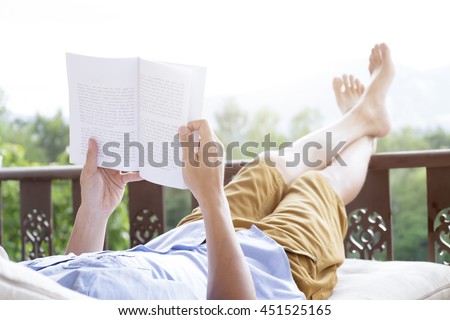 Young man reading a book lying on soft mattress in relaxing bed at terrace with green nature view. Fresh air in the morning of weekend or free day. Relax or education background idea. Selective focus.