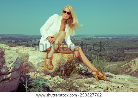 Blonde beautiful woman in white jacket on the rocks. Summer day. Hot weather. Fashion style. Fashion industry. Horizon. Summer wind