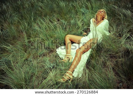 Blonde woman in sunshine rays under the sun on green grass, long legs tan. Summer day. Hot weather. Fashion industry. Field. Fashion style. Long legs woman. Lay in green grass. Under the sun