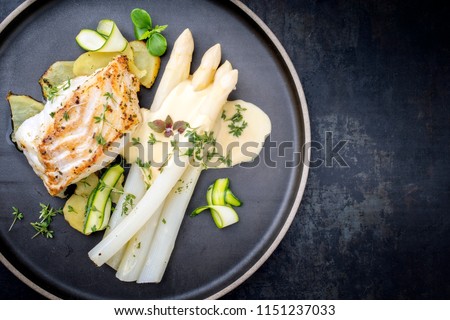 Modern German fried cod fish filet with white asparagus in hollandaise sauce with roast potatoes and sliced zucchini as top view on a plate with copy space right