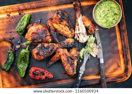 Traditional Caribbean barbecue jerk chicken wings and drumsticks with chimichurri sauce, jalapeno and poblano chili as top view on an old wooden cutting board