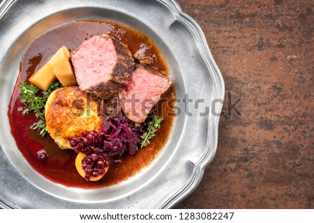 Traditional saddle of venison with fried mashed potatoes and red cabbage in game red wine sauce as top view on a pewter plate with copy space right