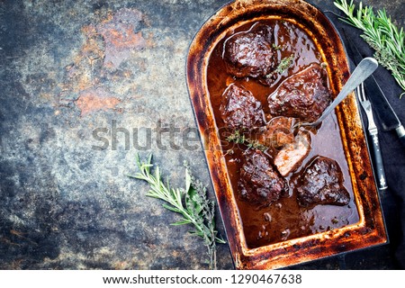 Traditional German braised veal cheeks in brown red wine sauce with mushroom and onions as top view in a stewpot with copy space left