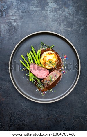 Traditional barbecue aged venison backstrap roast with green asparagus, fried mashed potatoes and herbs in brown red wine sauce as top view on a modern design plate with copy space