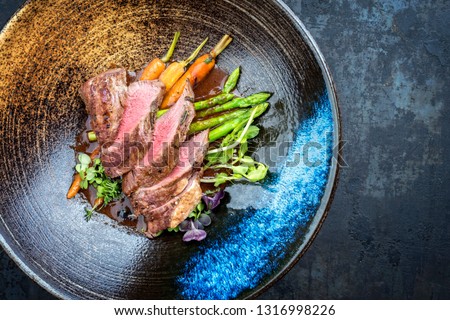 Traditional barbecue aged venison backstrap roast with green asparagus, carrots and herbs in brown red wine sauce as top view on a modern design bowl with copy space right