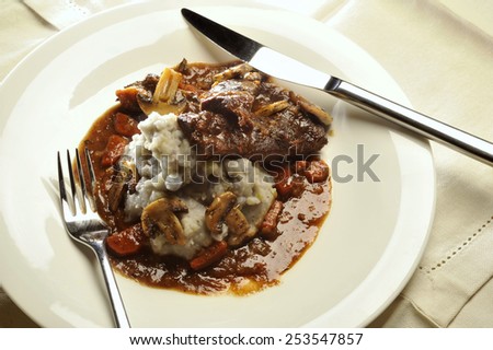 Beef cheeks in sauce with mushrooms and vegetables with mashed purple potatoes served on a white plate.