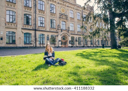 Beautiful, happy young student girl sitting on green grass near the backpack, bag under the tree near the campus, university, school, education. Summer, spring green park