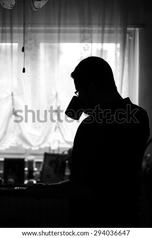 Man In Silhouette Drinking From Coffee Mug & Turning On The TV With A Remote In the Morning Against Bright Curtained Windows In The Living Room In Black & White Tone