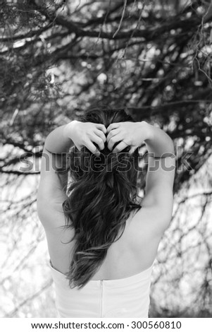 Portrait Of Caucasian Female Model Wearing Strapless White Dress Posing Near Forest Running Her Fingers Through Her Hair With Back Turned & Face Not Showing In Black & White